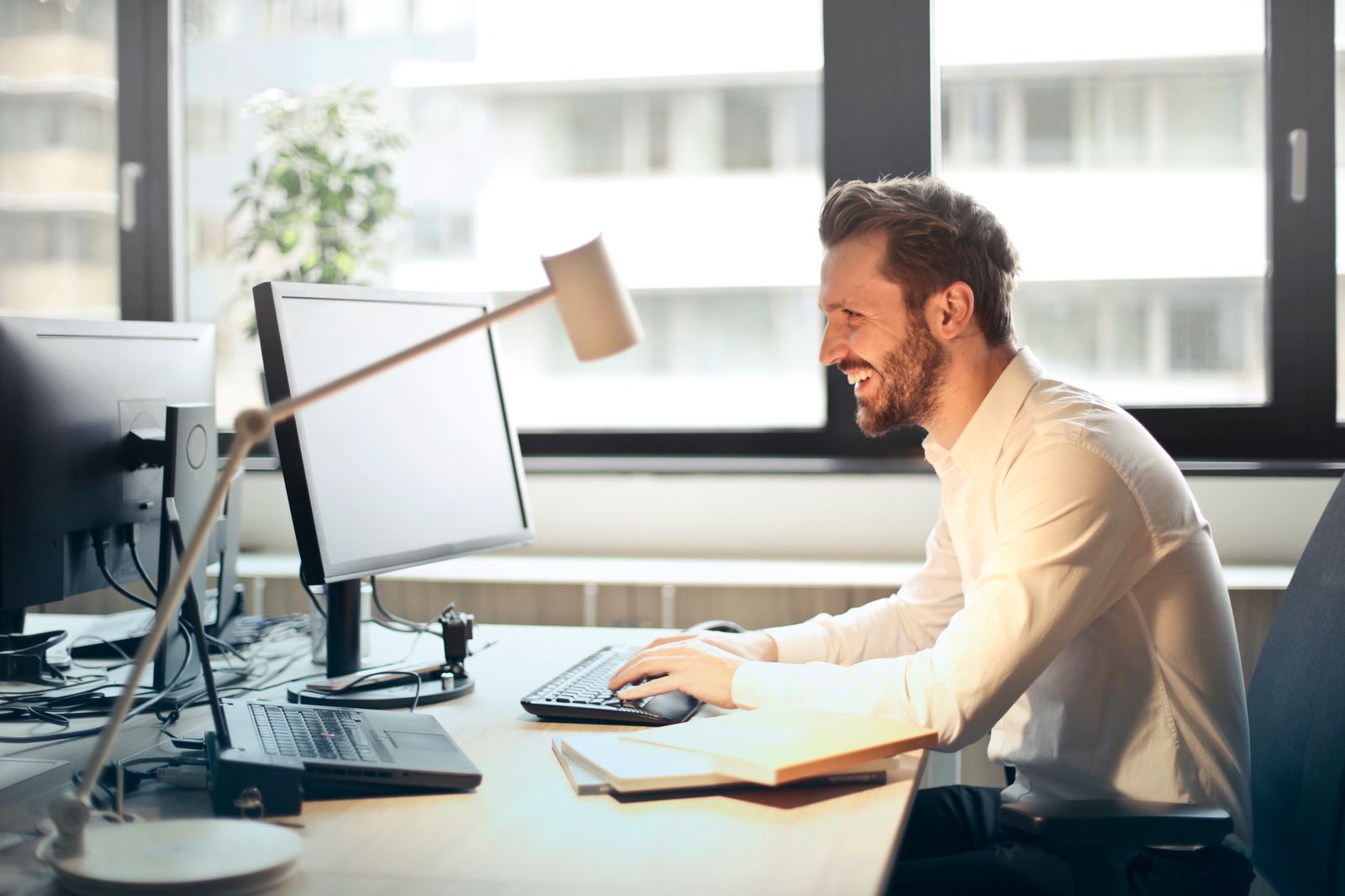 Homem de camisa branca trabalha sorrindo diante de monitor e teclado em um escritório iluminado.