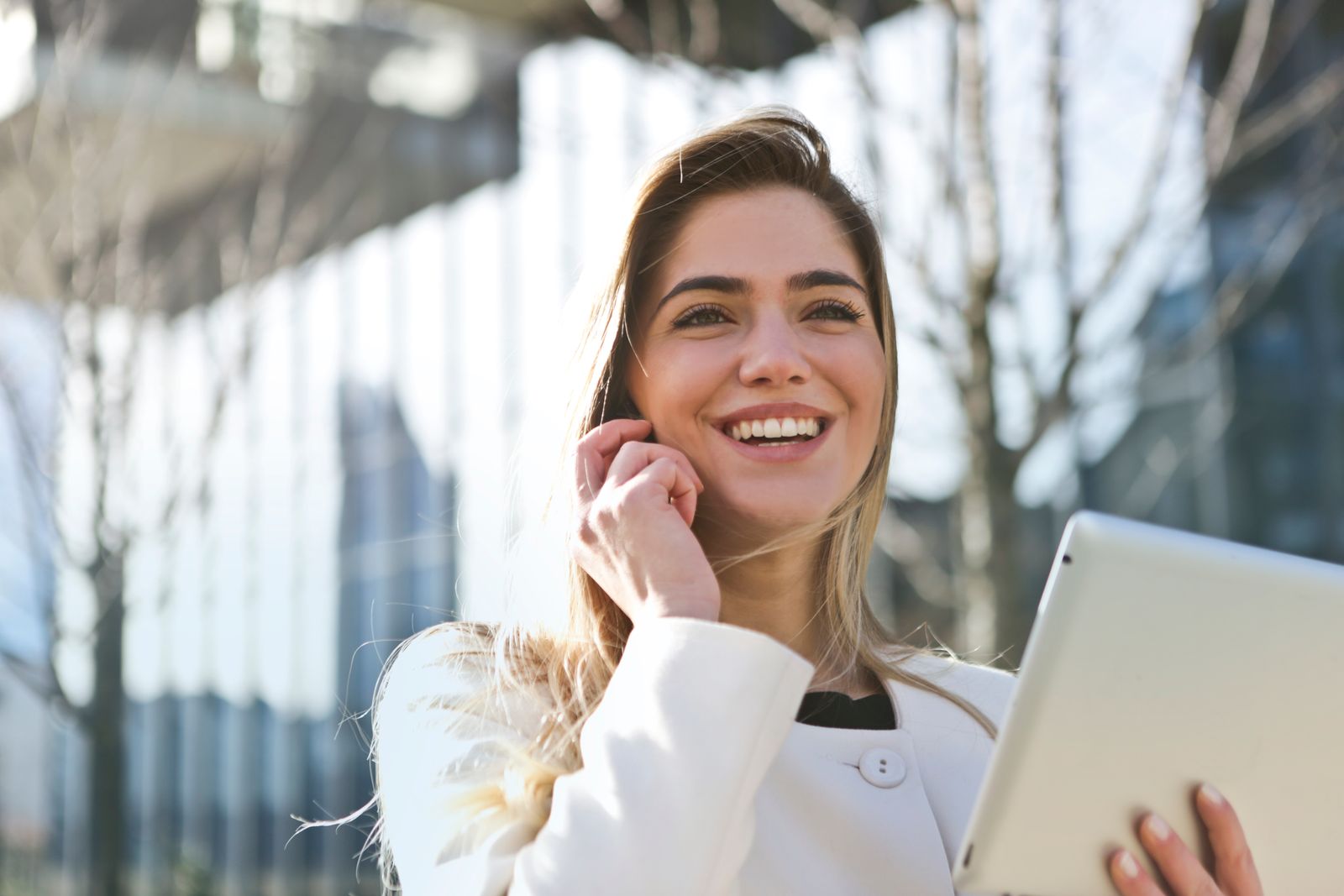 Mulher de blazer branco fala ao telefone segurando um tablet em frente a um prédio comercial.