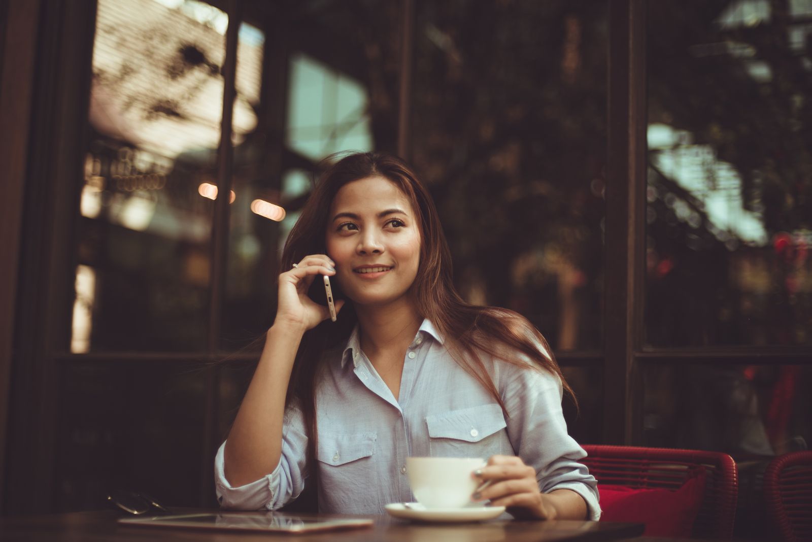 Mulher de camisa clara fala ao celular sentada à mesa de um café com xícara e tablet.